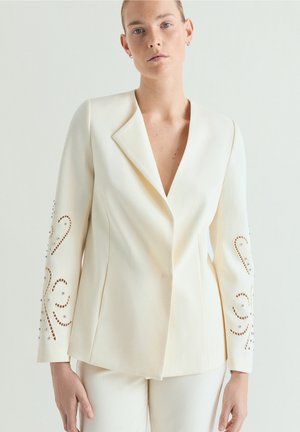 Vrouw draagt een off-white blazer met decoratieve uitsnijdingen en versieringen op de mouwen, gecombineerd met bijpassende pantalon, tegen een neutrale achtergrond.