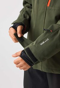 Grön utomhusjacka med justerbara ärmslut, utrustad med kardborrestängning och en ficka med dragkedja. Materialet har en slät textur med en matt yta.