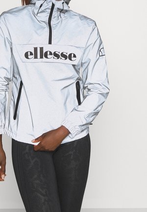 Personne portant une veste ellesse argentée avec des poches zippées noires et des leggings noirs texturés sur un fond clair uni.