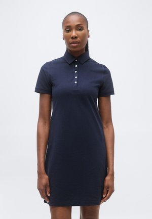 Vrouw die frontaal staat, gekleed in een donkerblauwe polo jurk met korte mouwen en een geknoopte kraag tegen een effen achtergrond.