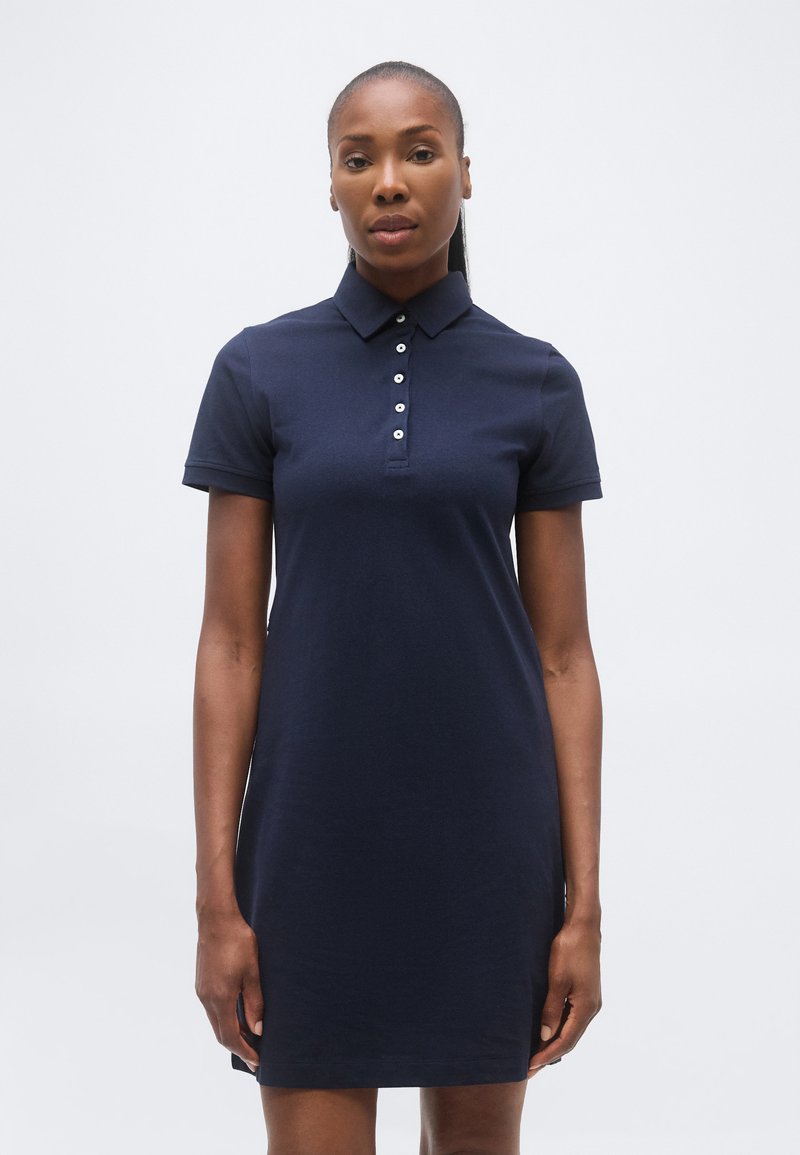 Γυναίκα που στέκεται με πρόσωπο προς τα εμπρός, φορώντας ένα σκούρο μπλε κοντομάνικο φόρεμα πόλο με γιακά με κουμπιά, μπροστά σε απλό φόντο.