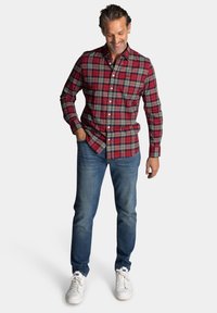 Rood en groen geruit button-up shirt met lange mouwen, gedragen met blauwe jeans en witte sneakers. Het shirt heeft een normale kraag en een borstzakje.