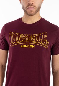 Tamsiai raudona medvilnė marškinėliai trumpomis rankovėmis su ryškiu geltonu "LONSDALE LONDON" logotipu per krūtinę.