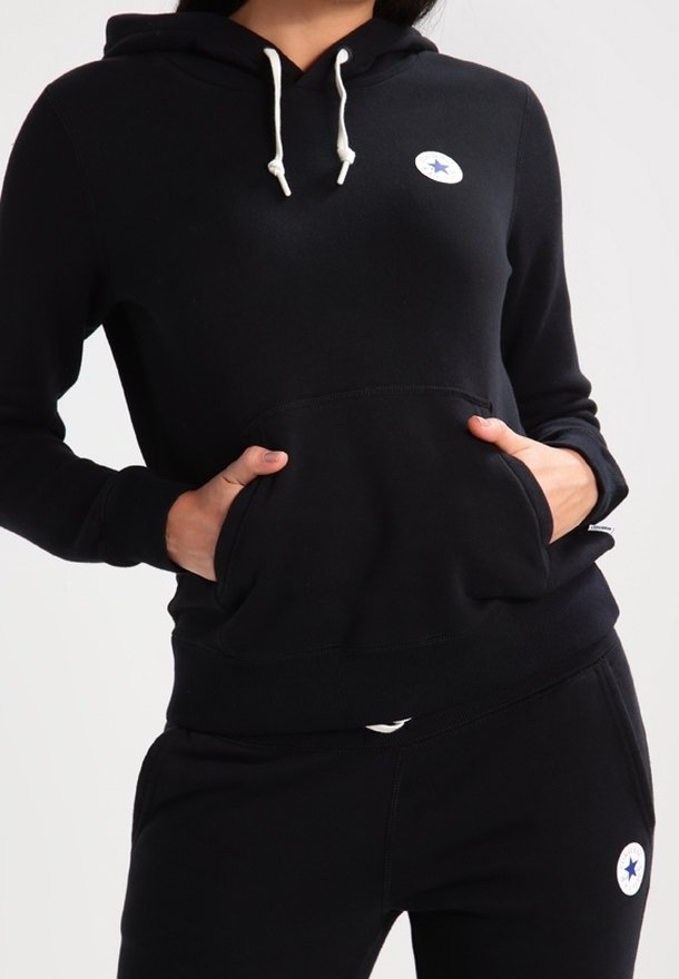 Personne portant un sweat à capuche noir et un pantalon avec des cordons blancs, tous deux avec de petits logos circulaires blancs avec des étoiles bleues.