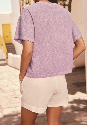 Personne debout à l'extérieur, les mains dans les poches, portant une chemise lavande à manches courtes et un short blanc sur un chemin pavé ensoleillé.