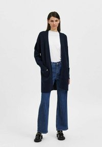 Cardigan blu navy realizzato in un tessuto morbido, con apertura frontale, maniche lunghe e due tasche laterali. Abbinato a jeans a zampa d'elefante a vita alta.