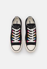 Zapatillas de lona negras con suela de goma blanca, que presentan ojales multicolores y cordones blancos. Logo notable en la lengüeta.