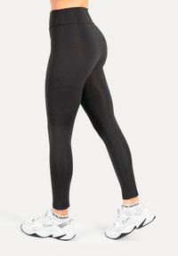 Leggings neri con vita alta, texture liscia e design aderente, abbinati a scarpe da ginnastica bianche.