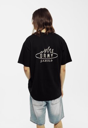 Camiseta negra de algodón con un gráfico blanco en la parte posterior que dice "GRMY" en letras estilizadas, combinada con pantalones cortos de denim claros.