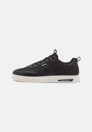 Zapatillas deportivas negras con una parte superior texturizada, suela blanca y un branding sutil. Presentan un diseño con cordones y una tira en la parte trasera para facilitar su uso.
