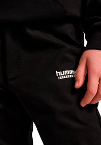 Sorte joggingbukser lavet af et blødt stof med et markant hvidt "hummel" logo på siden. Designet har en afslappet pasform og elastisk talje.