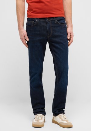 Mann trägt dunkelblaue Jeans, rotes Hemd und beige Sneaker, steht mit entspannten Händen an den Seiten vor einfarbigem Hintergrund.