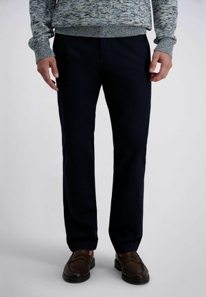 Mann trägt dunkelblaue Hose, braune Lederloafer und einen blau-grauen melierten Strickpullover, steht vor einem schlichten weißen Hintergrund.