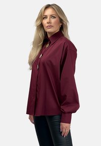 Burgundowa koszula z długim rękawem i wysokim kołnierzem, z zapięciem na guziki i metalicznymi akcentami. Gładki materiał o luźnym kroju.