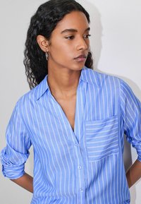 Light blue button-up shirt with narrow white stripes, featuring a chest pocket and rolled sleeves, made of smooth, breathable fabric.