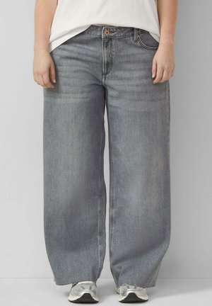 Person trägt locker sitzende graue Denim-Jeans, weiße Sneaker und ein weißes Oberteil, steht vor einem einfarbigen hellgrauen Hintergrund.