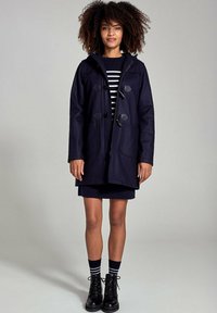 Marineblauwe wollen duffeljas met togglesluiting, twee voorkovacnten en een capuchon, gecombineerd met een gestreepte top en zwarte veterschoenen.