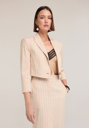 Kvinde iført en beige mørkeblåstribet cropped blazer og matchende nederdel med en silke beige top med sort meshdetalje, accessoriseret med perleøreringe.