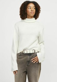 Pull en maille écru à col roulé avec une texture côtelée et des manches amples, associé à un pantalon marron taille haute et une ceinture claire.
