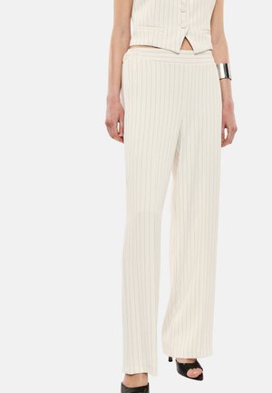 Donna con pantaloni crema a gamba larga a righe sottili, gilet smanicato abbinato, bracciale a polsino argentato e tacchi neri open toe, in piedi davanti a uno sfondo bianco.