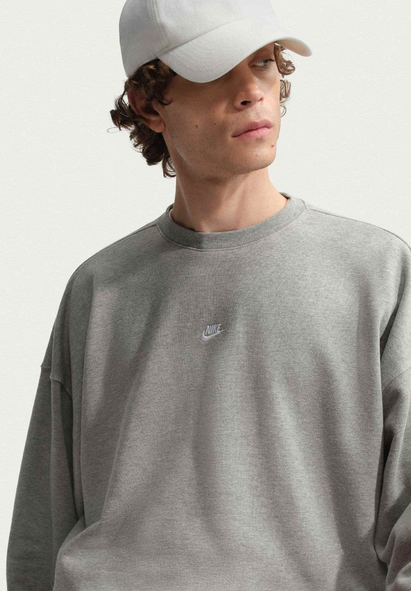Jeune homme aux cheveux bouclés portant un sweat-shirt Nike gris clair et une casquette de baseball blanche, regardant sur le côté sur un fond uni.