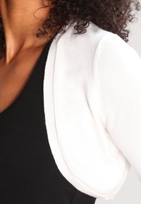 Biały krótki kardigan z dzianiny, z zaokrąglonym brzegiem i wszywanymi rękawami, noszony na czarnym topie.