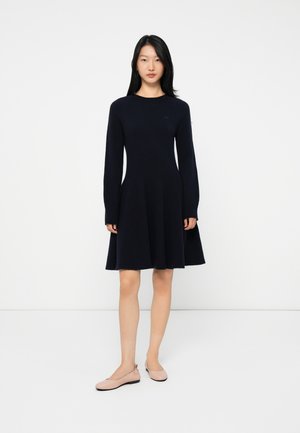 Femme se tenant contre un fond blanc, portant une robe bleue marine à manches longues et longueur genoux, ainsi que des ballerines rose pâle.