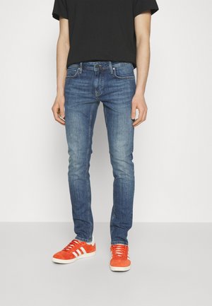Person, die eine Slim Fit blaue Jeans, ein schwarzes T-Shirt und leuchtend orange Sneakers mit weißen Streifen trägt, steht vor einem einfarbigen Hintergrund.