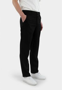 Schwarze maßgeschneiderte Hose mit geradem Schnitt, ausgestattet mit Seitentaschen und einem Reißverschluss, getragen mit einem weißen T-Shirt und weißen Sneakers.