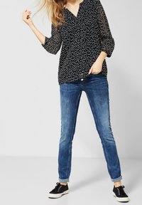 Blouse noire transparente à pois blancs, décolleté en V et manches trois-quarts ; associée à un jean en denim bleu et des baskets noires.
