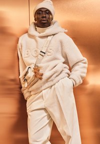 Hanorac oversize crem din fleece cu „BOSS” embossat, asortat cu pantaloni albi, cu talie înaltă, și o geantă mică crem, decorată cu un charm în formă de ursuleț.