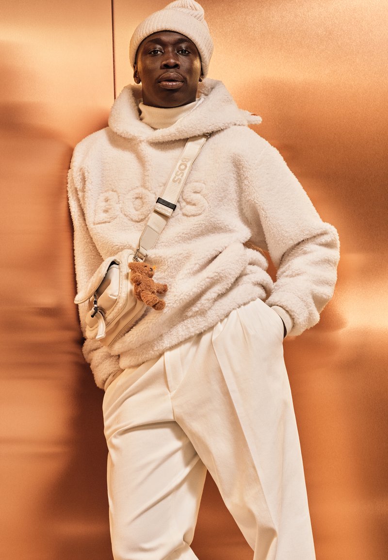 Hanorac oversize crem din fleece cu „BOSS” embossat, asortat cu pantaloni albi, cu talie înaltă, și o geantă mică crem, decorată cu un charm în formă de ursuleț.