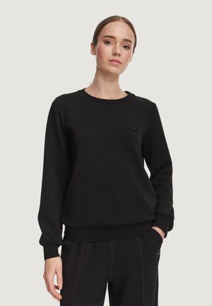 Giovane donna che indossa una felpa nera a maniche lunghe e pantaloni neri abbinati, con le mani nelle tasche, in piedi davanti a uno sfondo semplice.