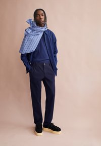 Marineblauw shirt en broek met een lichtblauwe gestreepte sjaal over één schouder gedrapeerd. Zwarte plateau schoenen met een beige zool. Gladde stofstructuur.