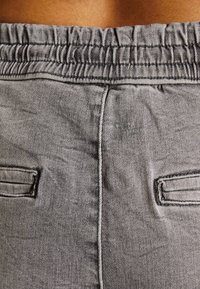 Gros plan d’un pantalon en tissu gris avec taille élastique et deux poches passepoilées à l’arrière, montrant la texture et les détails des coutures.