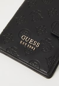 Black leather wallet with embossed logo pattern, featuring a gold "GUESS EST 1981" emblem. Smooth texture, rectangular shape, and snap closure.