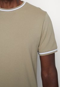 T-shirt beige à manches courtes en tissu texturé, avec un col rond en côte blanc et des bords de manches. Tissu lisse au design minimaliste.