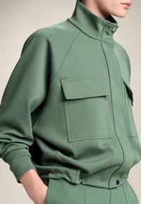 Felpa verde con colletto alto, due tasche frontali, tessuto a trama e design aderente. Presenta una chiusura a scatto all'escursione.