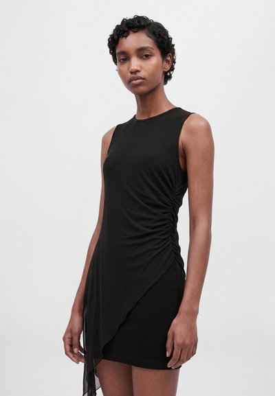 Un mannequin aux cheveux courts et bouclés se tient devant un fond uni, vêtu d’une robe noire sans manches, froncée sur les côtés et ornée d’un panneau drapé transparent.