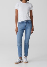 Personne portant un t-shirt blanc uni, un jean slim bleu clair et des baskets blanches avec des rayures bleu marine, debout devant un fond clair uni.