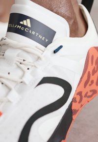 White athletic shoe featuring a black logo and laces, with an orange patterned sole and blue accent. Smooth material and distinct design details.