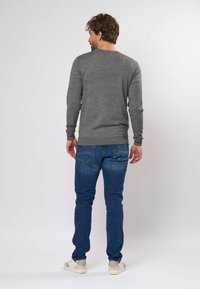 Mies seisoo selin harmaa pitkähihaainen villapaita, siniset farkut ja valkoiset tennarit päällään, yksiväristä vaaleaa taustaa vasten.