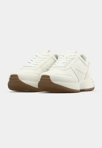 Λευκά αθλητικά παπούτσια με υφή στην επάνω επιφάνεια, επίπεδα κορδόνια και καφέ καουτσούκ σόλα με ζιγκ ζαγκ σχέδια για καλύτερη πρόσφυση. Στοιχεία μινιμαλιστικού σχεδιασμού.