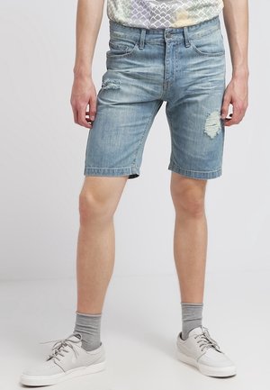 Personne portant un short en jean bleu clair usé, des chaussettes grises et des baskets blanches, debout, les jambes légèrement écartées.