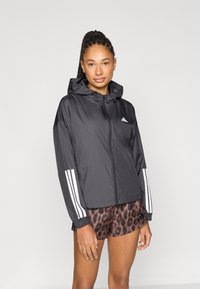 Sort lynlås-windbreaker med hætte, med tre hvide striber på hver ærme. Båret over brune leopardmønstrede shorts. Glat tekstur.