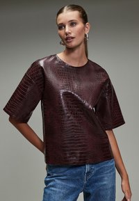 Burgundy, glossy faux leather top with a crocodile texture, short sleeves, and a round neckline, paired with denim jeans.