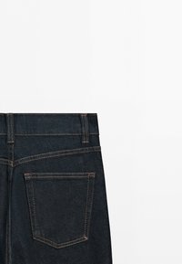 Back view of dark blue denim jeans showing waistband, belt loops, and one back pocket with orange stitching on white background.