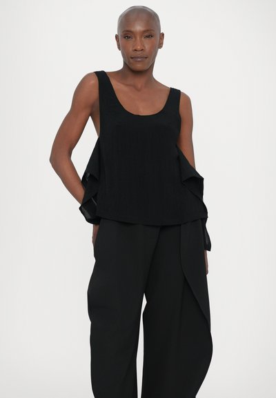 Black sleeveless top with a scoop neckline, flowing side drapes, textured fabric, paired with black wide-leg trousers.