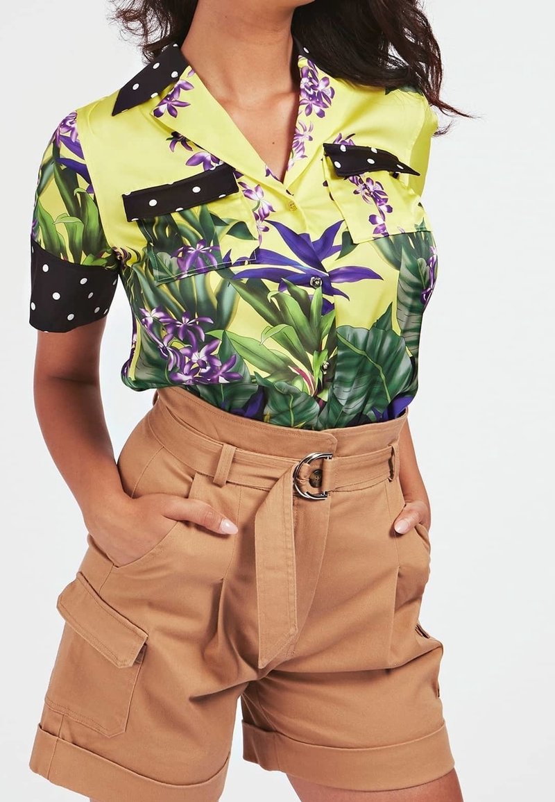 Femme portant un chemisier à manches courtes à motif floral jaune et vert avec des accents à pois noirs, et un short taille haute beige ceinturé avec des poches.