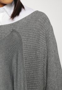 Grijze gebreide trui met een geribbelde textuur, brede halslijn en losse pasvorm. Wordt gedragen over een wit overhemd met kraag, wat het contrast tussen materialen versterkt.